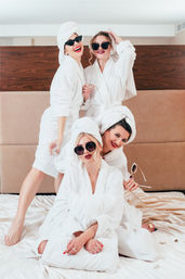 Four women in white bathrobes and towel turbans wearing sunglasses and sipping champagne while posing playfully on a hotel bed for a girls' spa day