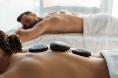 Couples hot-stone massage in a bright spa room — smooth black basalt stones lined along a bare back while a partner relaxes on a nearby table for relaxation and wellness