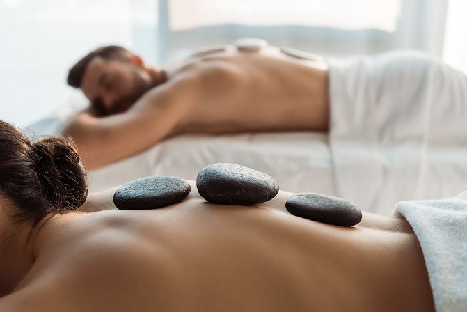 Couples hot-stone massage in a bright spa room — smooth black basalt stones lined along a bare back while a partner relaxes on a nearby table for relaxation and wellness