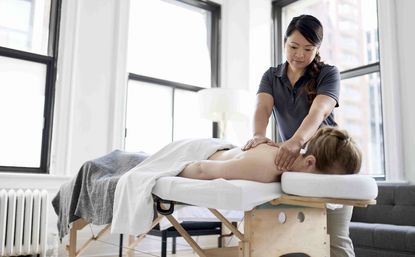 Massage therapist giving a back and shoulder massage to a client face down on a massage table in a bright urban wellness studio.