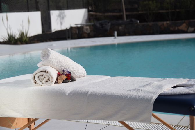 Sunlit poolside spa massage table draped in white towels with two rolled towels and small tropical flowers, turquoise outdoor pool in the background