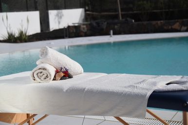Sunlit poolside massage table draped in white towels with two rolled towels, decorative stones and small flowers beside a turquoise outdoor swimming pool.