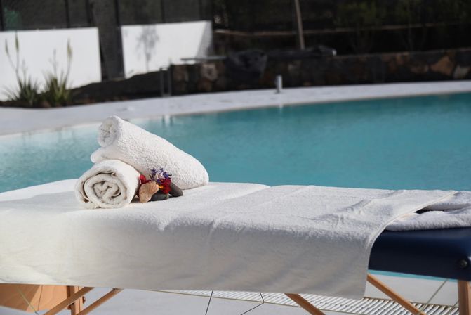 Sunlit poolside massage table draped in white towels with two rolled towels, decorative stones and small flowers beside a turquoise outdoor swimming pool.