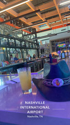 Nashville International Airport terminal bar with illuminated counter, a cocktail with straw and a cowboy hat on the bar, patrons, TVs and modern wood-grid ceiling