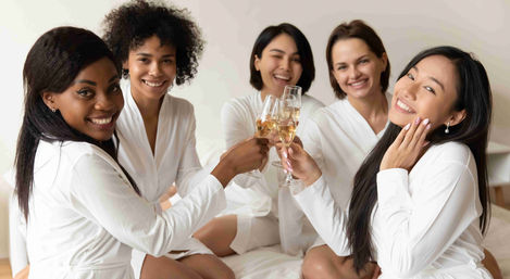 Five diverse women in white robes smiling and clinking champagne glasses on a bed — relaxed indoor spa-party or bachelorette-style celebration.