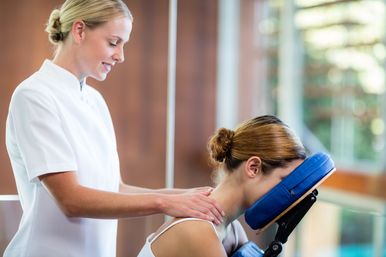 Smiling therapist gives a seated neck and shoulder massage with a blue face cradle in a bright wellness spa
