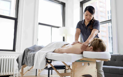 Massage therapist performing a relaxing back and shoulder massage on a client lying face down on a massage table in a bright, sunlit city studio.