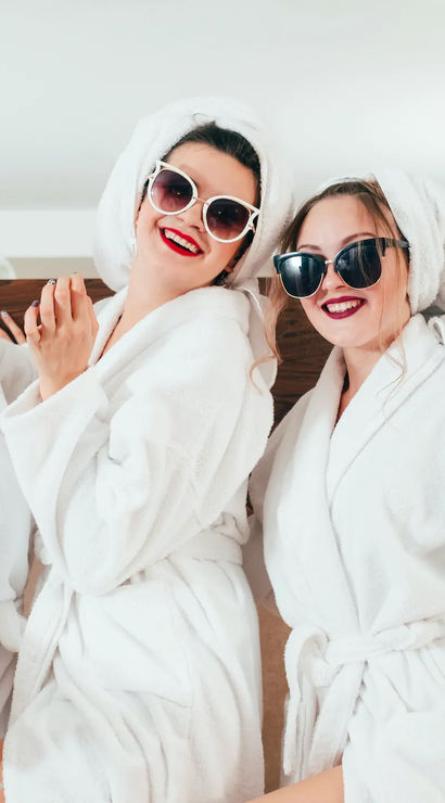 Three women in white bathrobes with towel turbans and sunglasses, red lipstick, toasting with champagne in a hotel room by a wooden headboard and bedside lamp — playful spa getaway vibe.