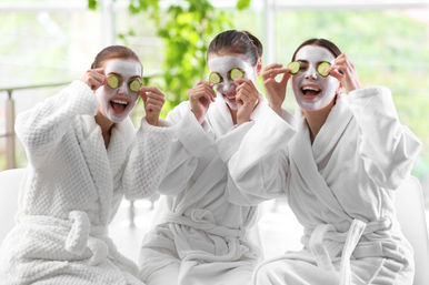 Three friends in plush white bathrobes wearing face masks and cucumber slices over their eyes, laughing during a bright indoor spa day.