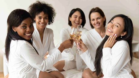 Group of five women in white robes smiling and clinking champagne glasses on a bed during a bridal shower or spa-style celebration.