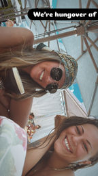 Two smiling friends lounging under beach umbrellas in a sunny poolside cabana — one wearing round sunglasses and a patterned headscarf holding a chocolate-covered ice cream bar; text overlay reads “We’re hungover af”.