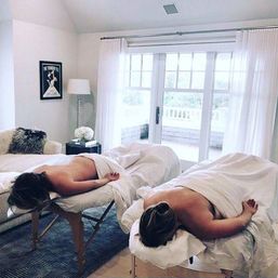 Two people lying face down on side-by-side massage tables covered in white sheets in a bright, sunlit spa-like room with sheer curtains, French doors to a balcony, a cozy sofa, lamp, and fresh flowers.