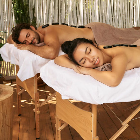 Serene outdoor couples hot-stone massage on a wooden deck with bamboo screen, two relaxed people lying on spa tables.