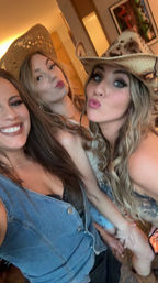 Three friends taking a playful indoor selfie at a western-themed party, wearing cowboy/cowgirl hats and casual denim, smiling and puckering lips.