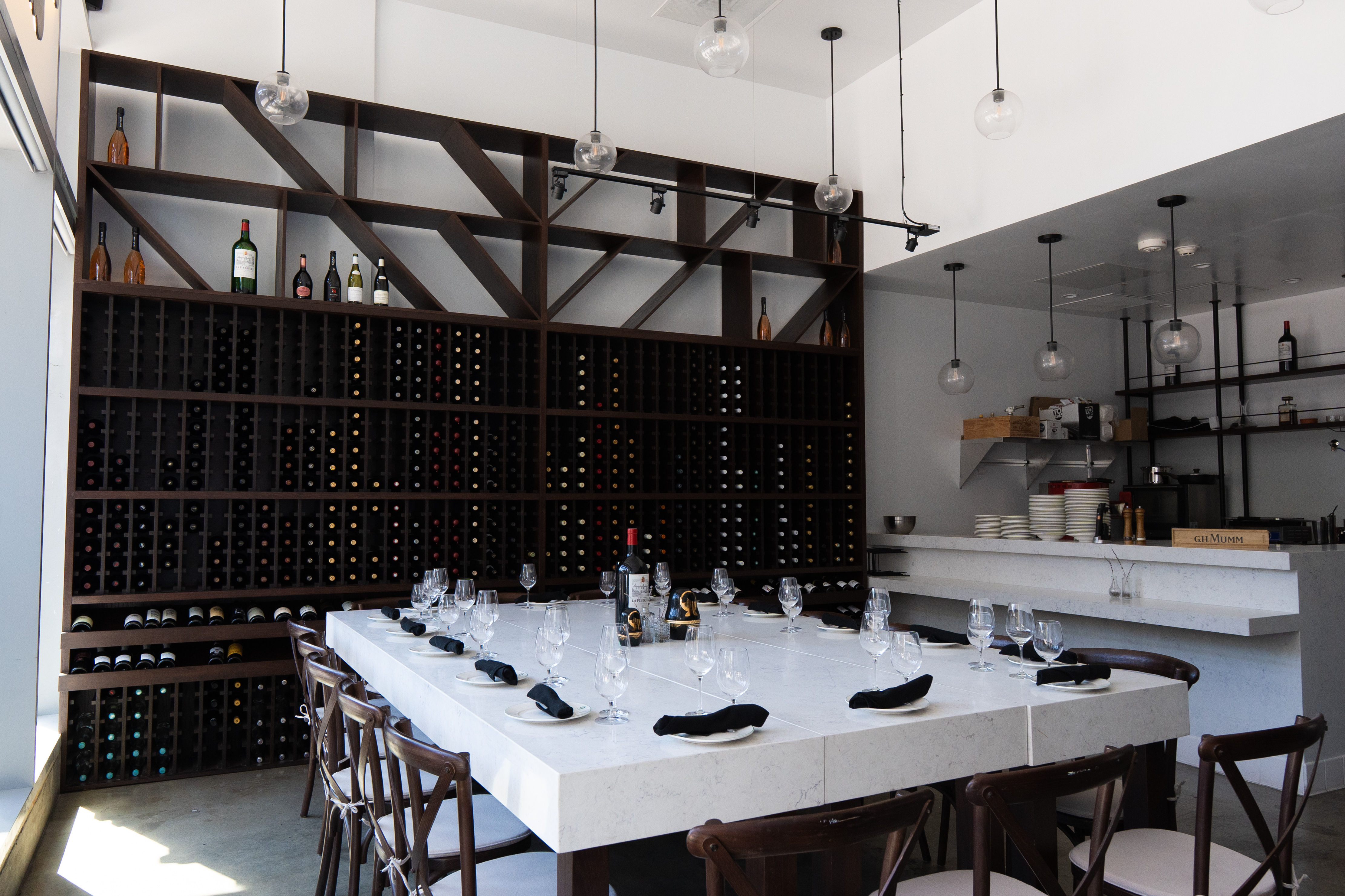 Modern wine bar interior with floor-to-ceiling wooden wine rack, marble communal table set with wine glasses and black napkins, and hanging globe pendant lights.