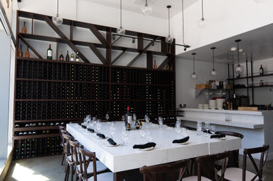 Modern wine bar interior with floor-to-ceiling wooden wine rack, marble communal table set with wine glasses and black napkins, and hanging globe pendant lights.
