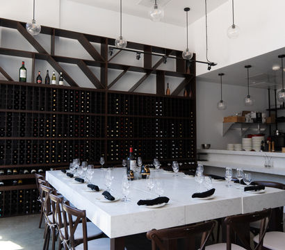 Modern wine bar interior with floor-to-ceiling wooden wine rack, marble communal table set with wine glasses and black napkins, and hanging globe pendant lights.