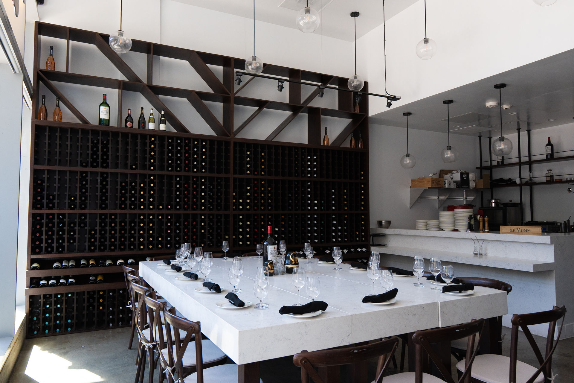 Modern wine bar interior with floor-to-ceiling wooden wine rack, marble communal table set with wine glasses and black napkins, and hanging globe pendant lights.