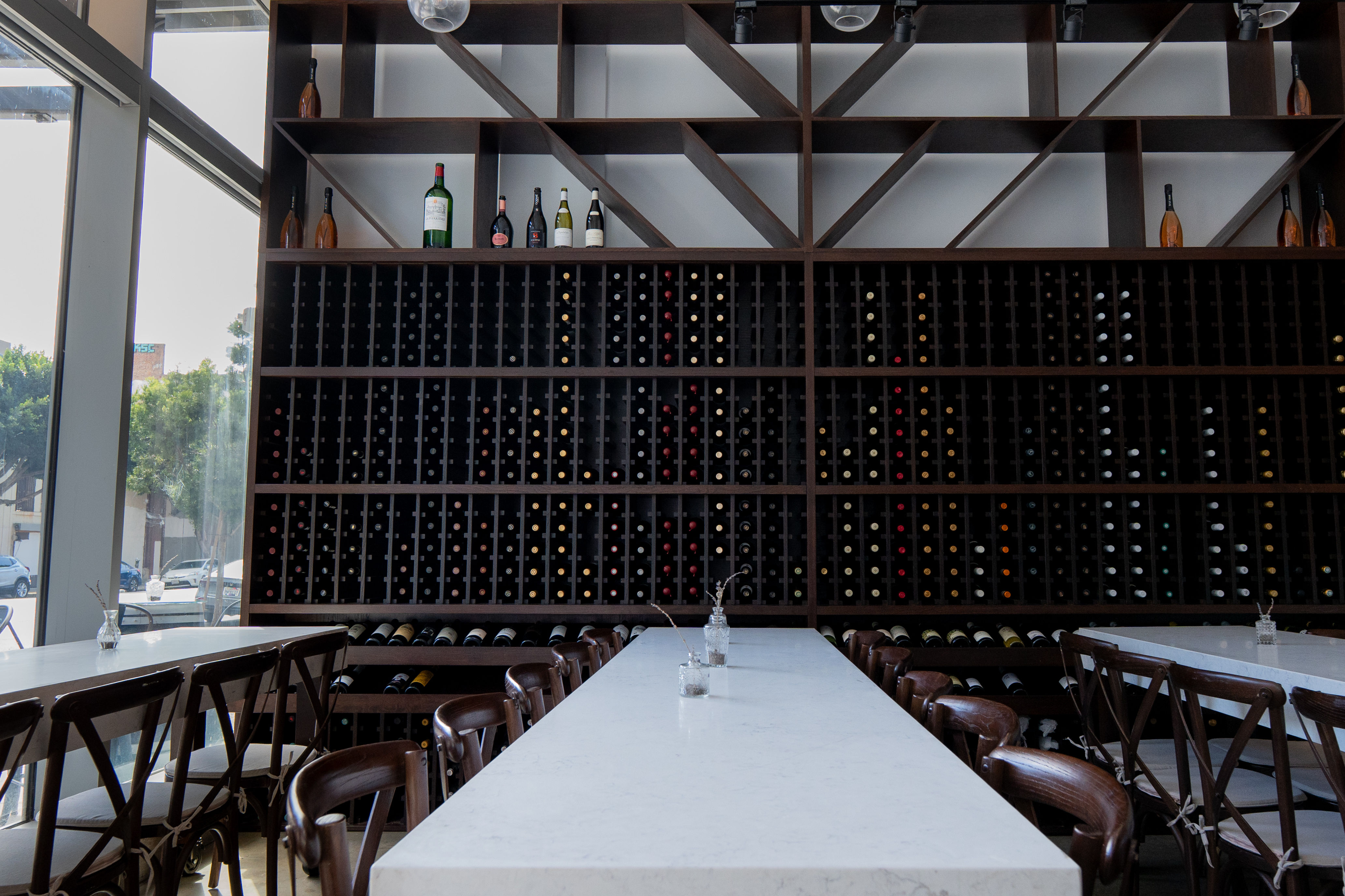 Sunlit modern downtown wine bar interior with a long white marble communal table, wooden chairs and a floor-to-ceiling dark wood wine rack filled with bottles and diagonal shelving