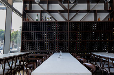 Sunlit modern downtown wine bar interior with a long white marble communal table, wooden chairs and a floor-to-ceiling dark wood wine rack filled with bottles and diagonal shelving