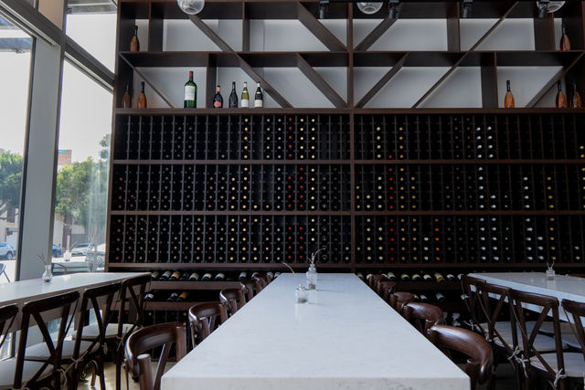 Sunlit modern downtown wine bar interior with a long white marble communal table, wooden chairs and a floor-to-ceiling dark wood wine rack filled with bottles and diagonal shelving