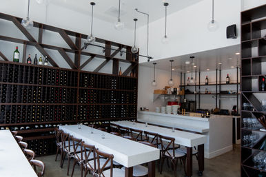 Modern wine bar interior with floor-to-ceiling wooden wine racks, long white communal tables, wooden chairs, marble bar counter and hanging globe pendant lights.