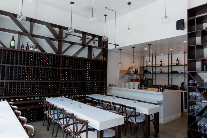 Modern wine bar interior with floor-to-ceiling wooden wine racks, long white communal tables, wooden chairs, marble bar counter and hanging globe pendant lights.