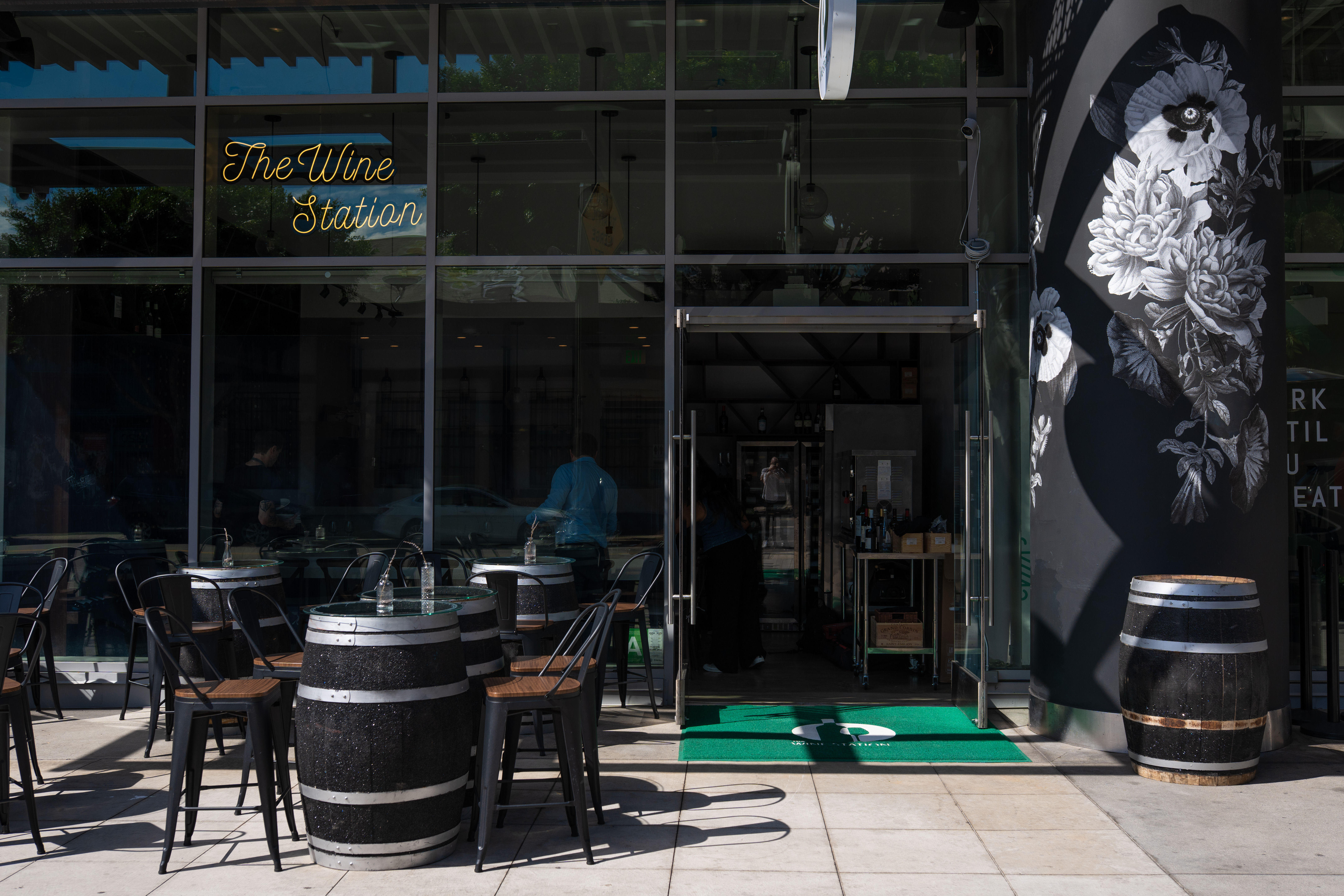 Glass-front wine bar storefront with outdoor patio of barrel-top tables and black metal chairs, neon script sign above the entrance, large black-and-white floral mural on the side wall, and an open glass door showing bottles inside.