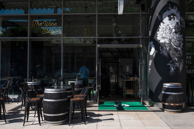 Glass-front wine bar storefront with outdoor patio of barrel-top tables and black metal chairs, neon script sign above the entrance, large black-and-white floral mural on the side wall, and an open glass door showing bottles inside.