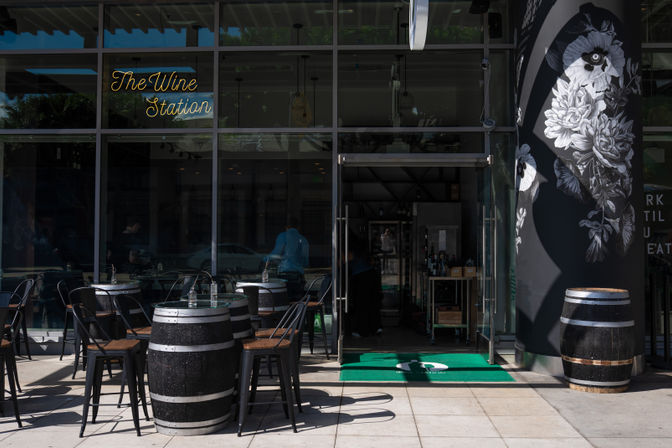 Glass-front wine bar storefront with outdoor patio of barrel-top tables and black metal chairs, neon script sign above the entrance, large black-and-white floral mural on the side wall, and an open glass door showing bottles inside.