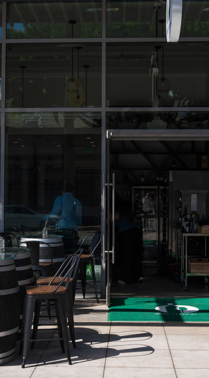 Glass-front wine bar storefront with outdoor patio of barrel-top tables and black metal chairs, neon script sign above the entrance, large black-and-white floral mural on the side wall, and an open glass door showing bottles inside.