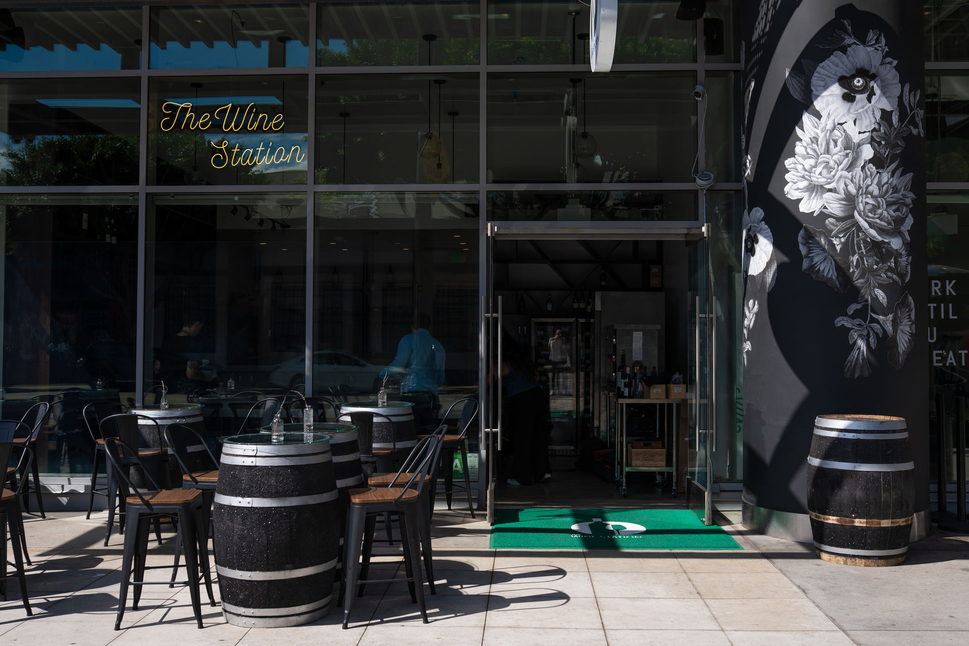 Glass-front wine bar storefront with outdoor patio of barrel-top tables and black metal chairs, neon script sign above the entrance, large black-and-white floral mural on the side wall, and an open glass door showing bottles inside.