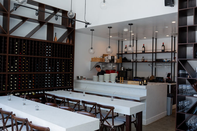 Modern wine bar interior with floor-to-ceiling wooden wine rack, white marble communal tables, globe pendant lights and a bar counter with stacked plates and staff prepping