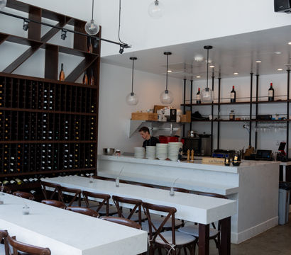 Modern wine bar interior with floor-to-ceiling wooden wine rack, white marble communal tables, globe pendant lights and a bar counter with stacked plates and staff prepping
