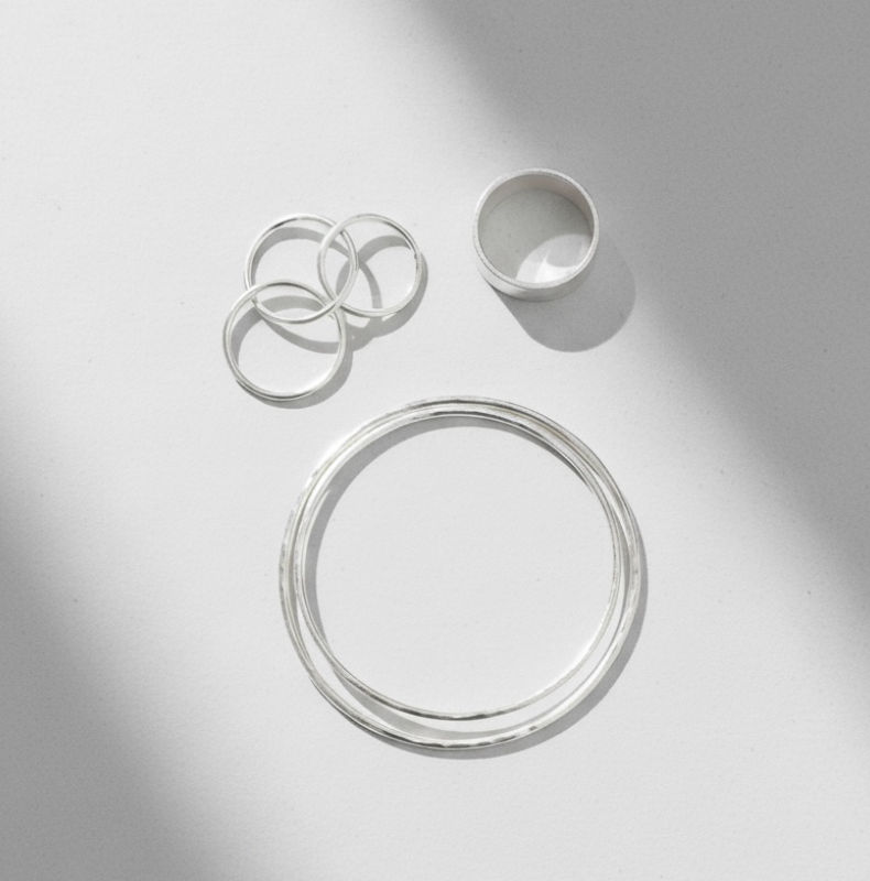 Minimalist flatlay of polished sterling silver bangles and three stacking rings on a white background with soft diagonal shadows