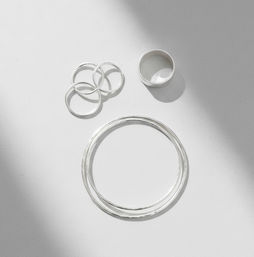 Minimalist flatlay of polished sterling silver bangles and three stacking rings on a white background with soft diagonal shadows