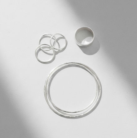 Minimalist flatlay of polished sterling silver bangles and three stacking rings on a white background with soft diagonal shadows