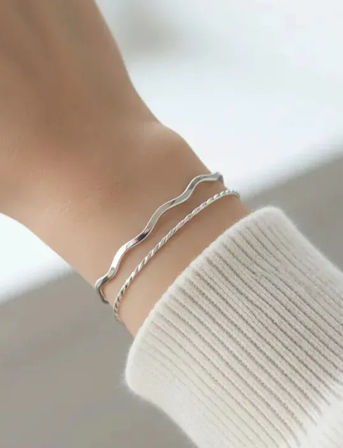 Close-up of a wrist wearing two delicate silver bracelets — a wavy band and a twisted rope chain — styled with a white ribbed sweater cuff, minimalist stacked jewelry