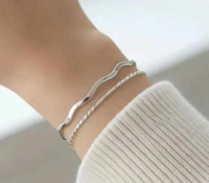 Close-up of a wrist wearing two delicate silver bracelets — a wavy band and a twisted rope chain — styled with a white ribbed sweater cuff, minimalist stacked jewelry