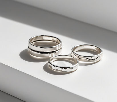 Close-up of three sterling silver stacking rings (smooth, hammered, wavy) on a white studio background with soft sunlight shadows — minimalist jewelry flatlay.