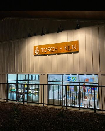 Nighttime exterior of a lit glassblowing studio with rust-colored metal sign, white board-and-batten facade and large windows showcasing colorful blown-glass art and an 'open' sign.