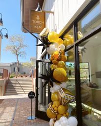 Sunlit outdoor storefront with glass door and open sign, entrance wrapped in a festive gold, white and black balloon garland with metallic chain accents and spiky star-shaped balloons.