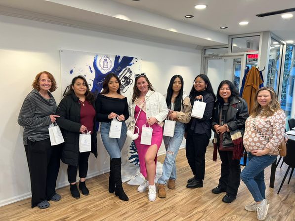 Smiling group of eight women holding white gift bags in a bright boutique-style studio with wood floors, wearing casual coats, jeans and a pink dress.