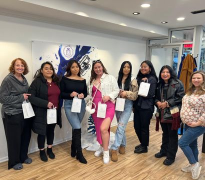 Smiling group of eight women holding white gift bags in a bright boutique-style studio with wood floors, wearing casual coats, jeans and a pink dress.