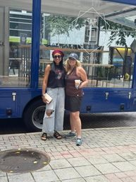 Two smiling women posing on a downtown sidewalk in front of a blue mobile shop with glass display; one wearing a red beret and sunglasses, the other holding an iced coffee.