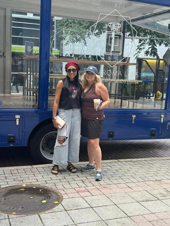 Two smiling women posing on a downtown sidewalk in front of a blue mobile shop with glass display; one wearing a red beret and sunglasses, the other holding an iced coffee.