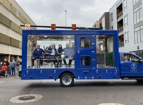Blue mobile display truck with glass-sided showroom and four people in lab coats inside, rolling through a downtown street parade past onlookers and mid-rise buildings under an overcast sky.