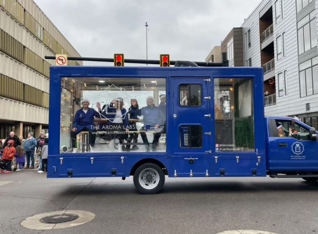 Blue mobile display truck with glass-sided showroom and four people in lab coats inside, rolling through a downtown street parade past onlookers and mid-rise buildings under an overcast sky.