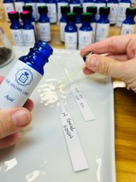 Cobalt blue fragrance bottle and pipette held over scent test strips labeled 'Emerald Hibiscus' and 'Açaí', with rows of similar blue bottles in the background — fragrance sampling station.