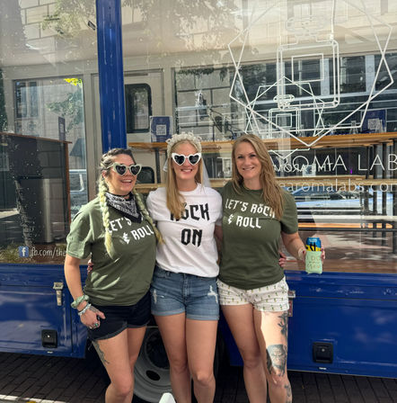 Three smiling women posing on a city sidewalk in front of a blue food truck — bachelorette in center with veil and heart sunglasses, friends in matching 'Let’s Rock & Roll' shirts.